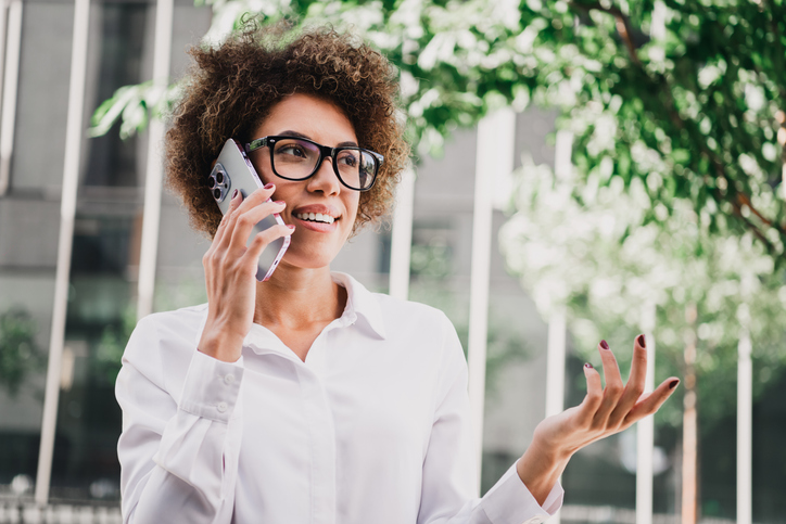 A confident young woman in a white shirt talks on her phone outdoors in a modern city setting with glass façades and trees, conveying business style urban lifestyle and professional poise.