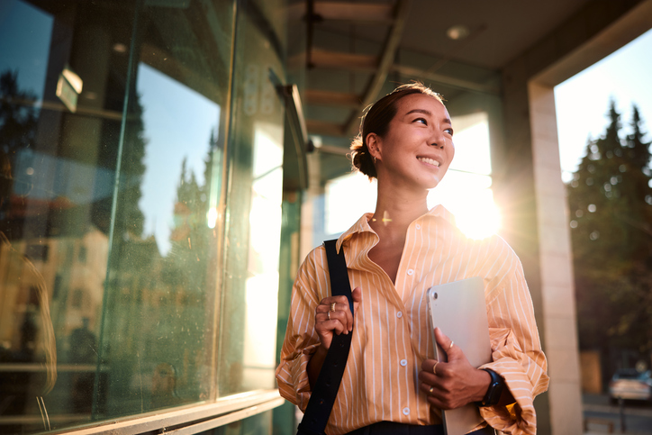 Young Asian businesswoman walking near office building holding tablet and smiling