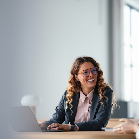 A friendly businesswoman in a gray blazer and pink shirt chats with a colleague across a desk in a bright, modern office. She smiles behind stylish glasses, highlighting teamwork and productive collaboration.
